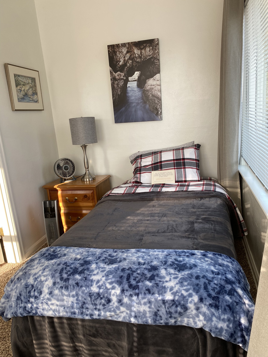 A bedroom with white walls, a wooden 3 drawer nightstand with a fan and lamp on it; two pictures hanging on the walls, a large window next to the bed with neutral curtains, brown shag carpet